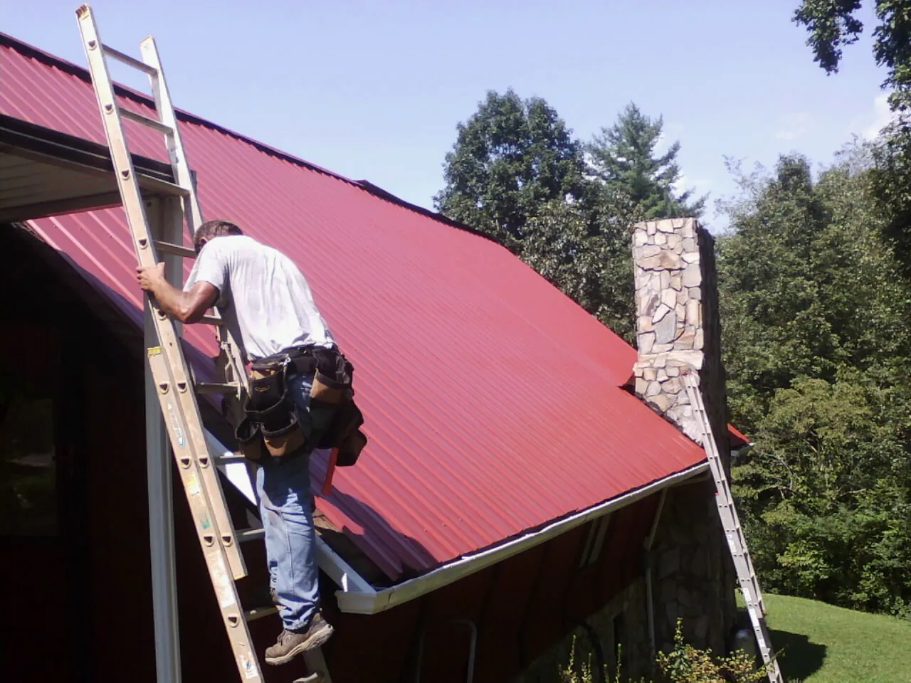 Roofer on a ladder beside a red metal roof and stone chimney — Soffit & Fascia Repair by Roofing Brothers in Dalton